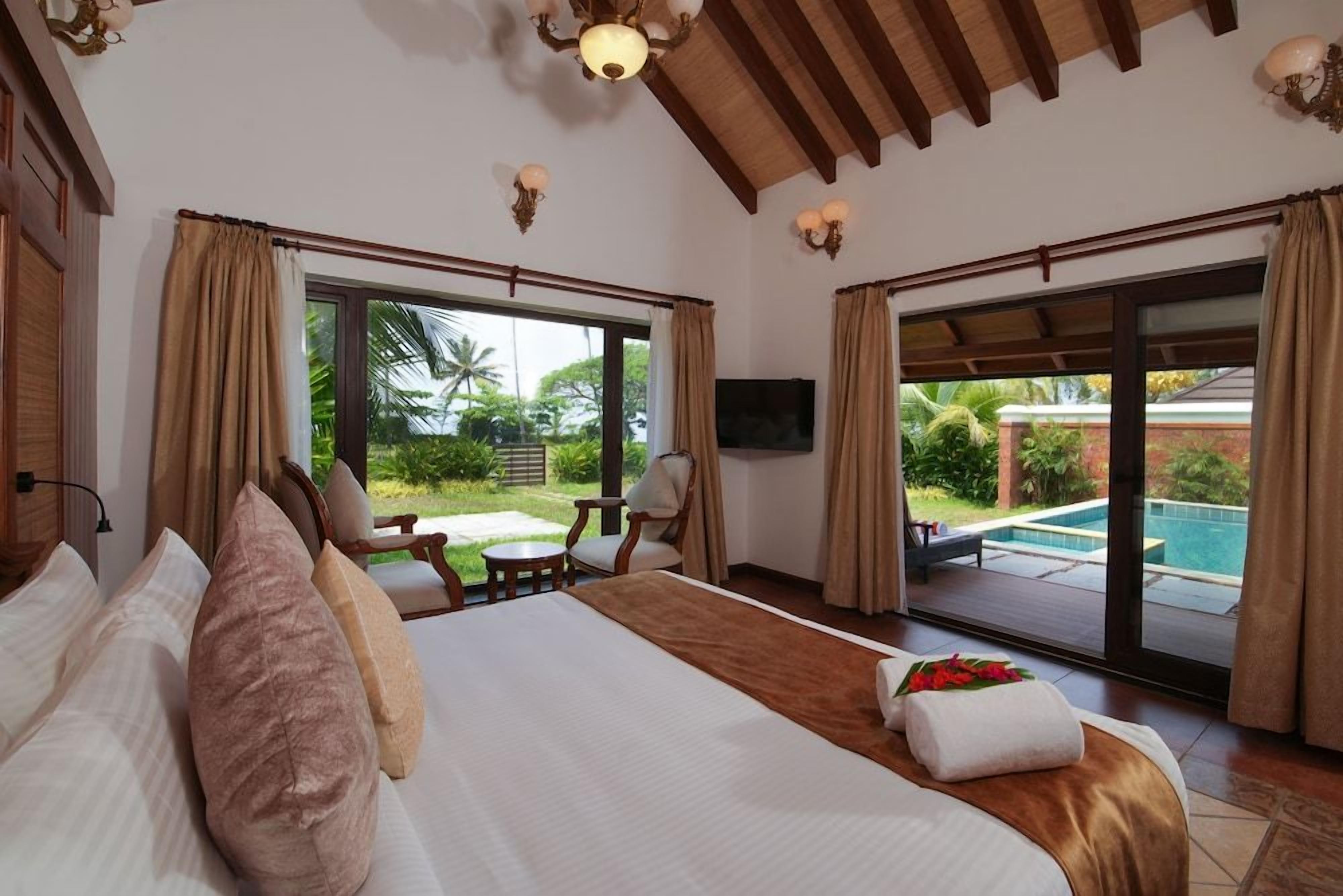 A hotel room with a large bed, a TV, a balcony with a pool, and a view of greenery.