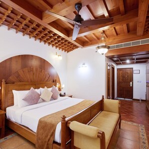 A wooden-framed bedroom with a bed, bedside tables, a bench, and a ceiling fan. - Gokulam Grand Resort and Spa Kumarakom (Kottayam)