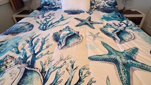 A bed with a sea-themed bedspread, a pillow, and a painting of a sunset on the wall.