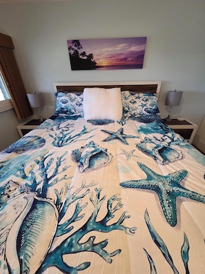 A bed with a sea-themed bedspread, a pillow, and a painting of a sunset on the wall.