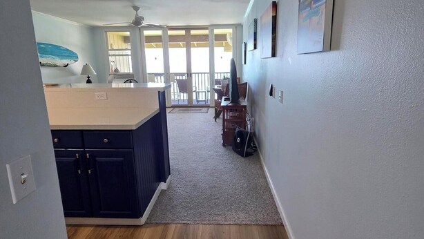 A spacious room with a wooden floor, a kitchen area with a counter and cabinets, a ceiling fan, and a view through large windows to an outdoor balcony.
