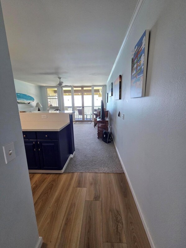 A spacious room with a wooden floor, a kitchen area with a counter and cabinets, a ceiling fan, and a view through large windows to an outdoor balcony.