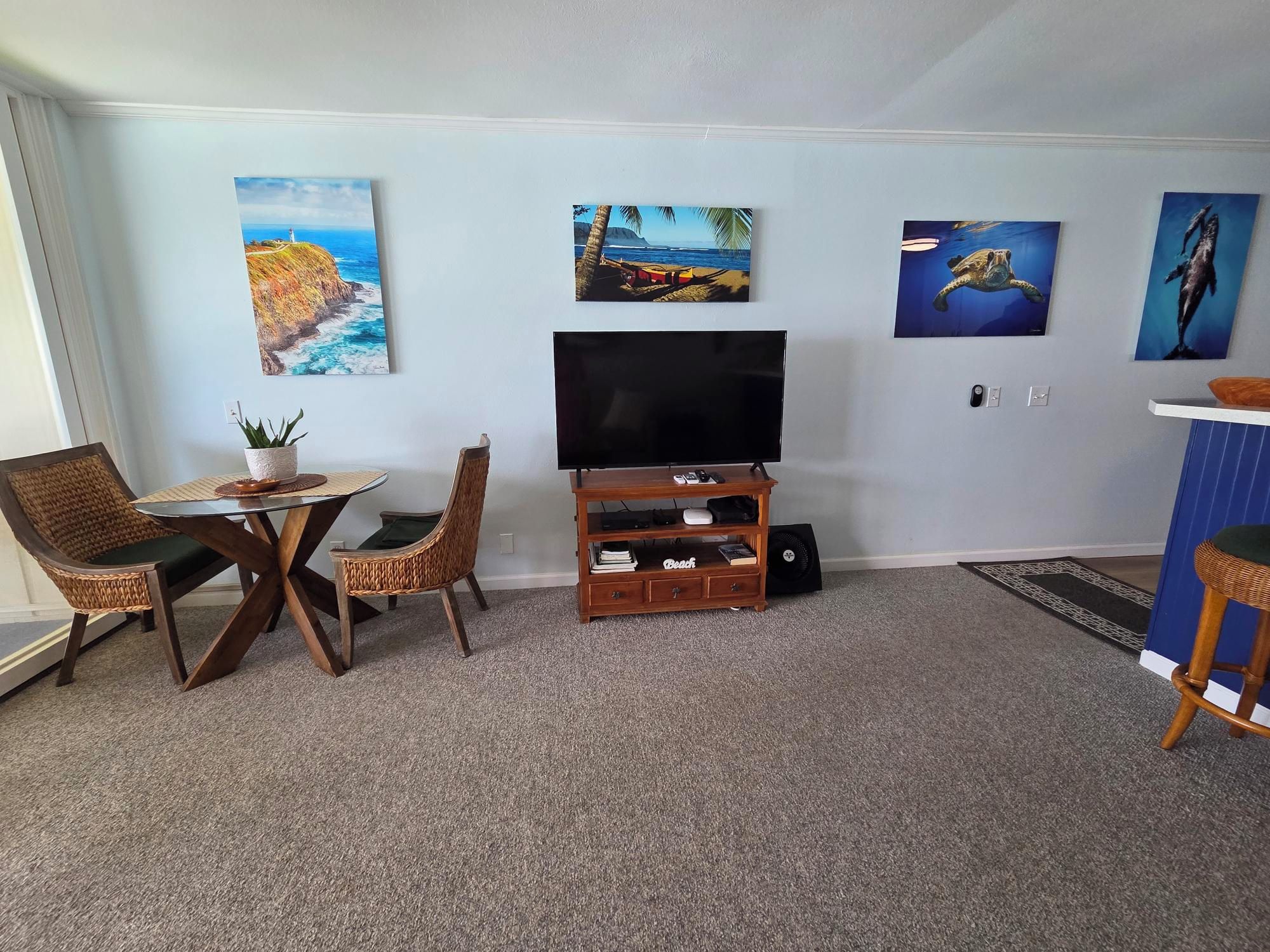 A living room with a flat-screen TV, wicker chairs, a small table with a plant, and artwork on the walls.