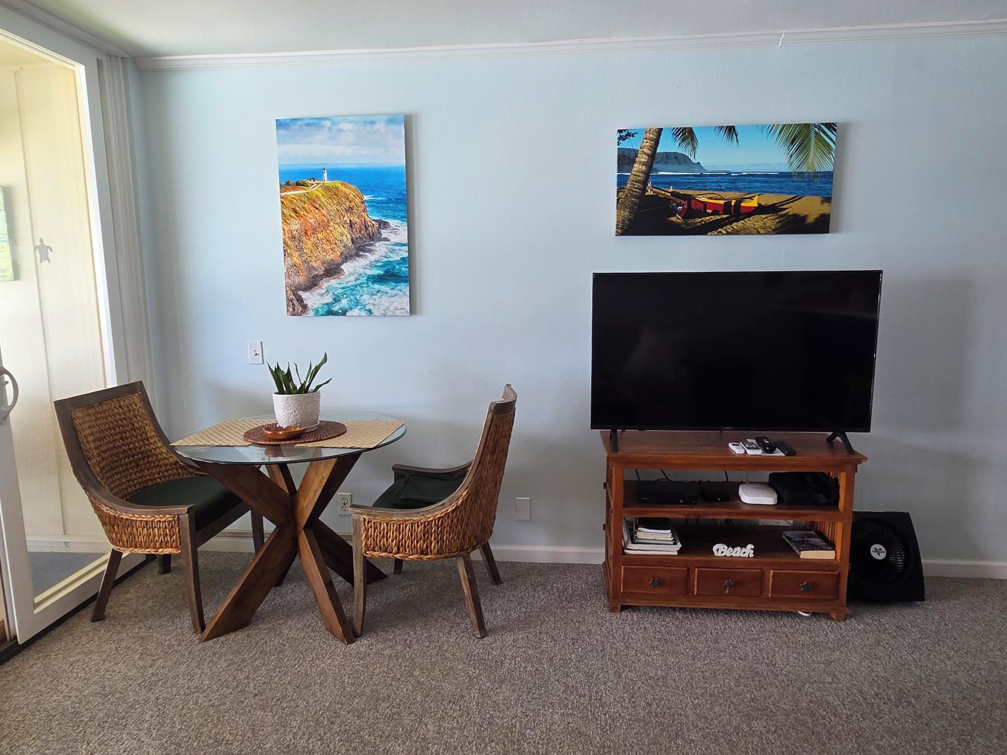 Een woonkamer met een televisie op een houten standaard, twee rieten fauteuils, een bijzettafeltje met een plant en twee schilderijen van kustgezichten.