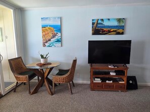 A living room with a television on a wooden stand, two wicker chairs, a small table with a plant, and two paintings of coastal scenes. - WOW! GORGEOUS TOP FLOOR DIRECT OCEANFRONT CONDO, TWO A/C UNITS, FREE PARKING! (Kapaʻa)