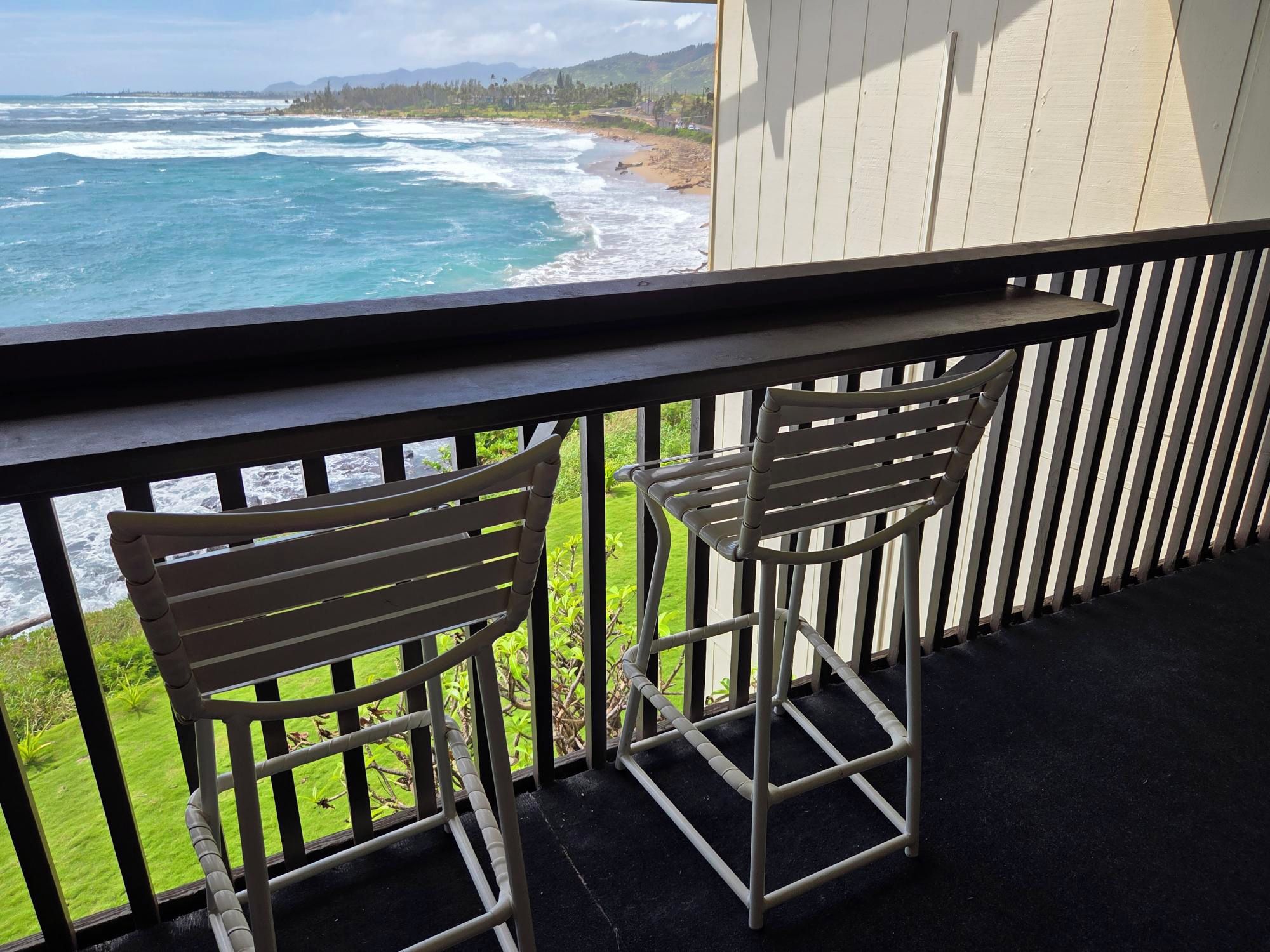 A balcony with two metal chairs, a view of the ocean, and a grassy area.