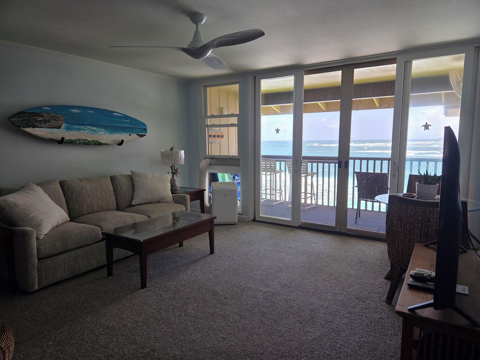 A living room with a sofa, a coffee table, a television, and a ceiling fan. There is a large window with a view of the ocean.