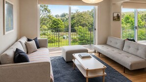 Uma sala de estar moderna com dois sofás, uma mesa de centro e vista para uma varanda com vegetação.