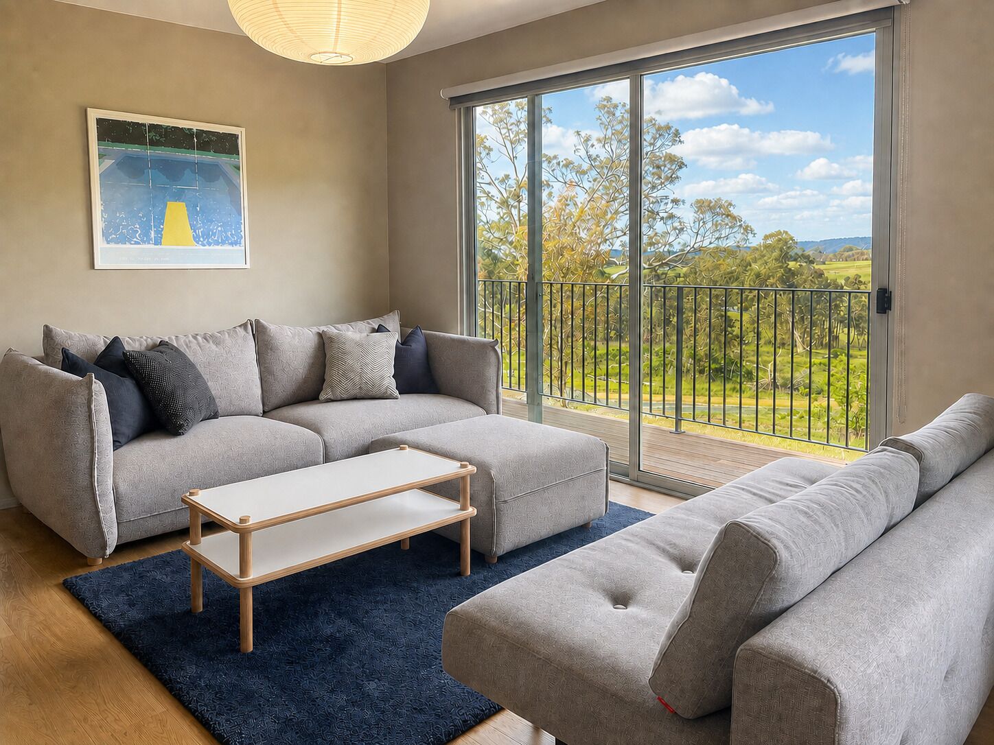 A modern living room with a grey sofa set, a coffee table, and a large sliding glass door leading to a balcony with a view of greenery.