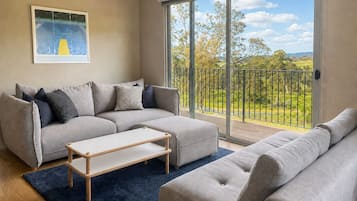 A modern living room with a grey sofa set, a coffee table, and a large sliding glass door leading to a balcony with a view of greenery.