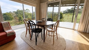 Uma sala de jantar com mesa redonda, cadeiras, um sofá vermelho e vista para um jardim através de grandes janelas.