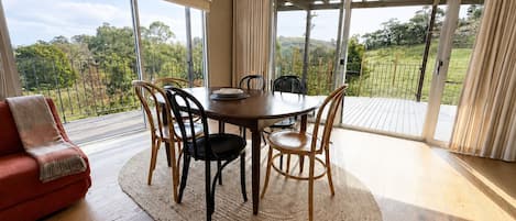 Comedor con mesa redonda, sillas, sofá rojo y vistas a un jardín a través de grandes ventanales.