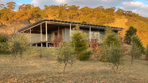 Um edifício térreo com varanda coberta e telhado plano, cercado por grama seca e árvores esparsas.