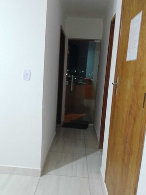 A narrow hallway with a wooden door on the right, a white wall with a light switch, and a view of a balcony with lights outside.