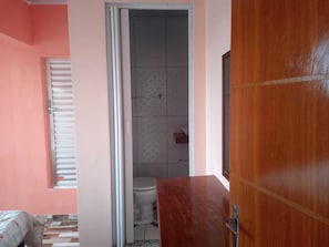 Una camera da letto con un letto, una cassettiera in legno, una finestra con persiane e un ingresso che conduce a un bagno con il water e una doccia.