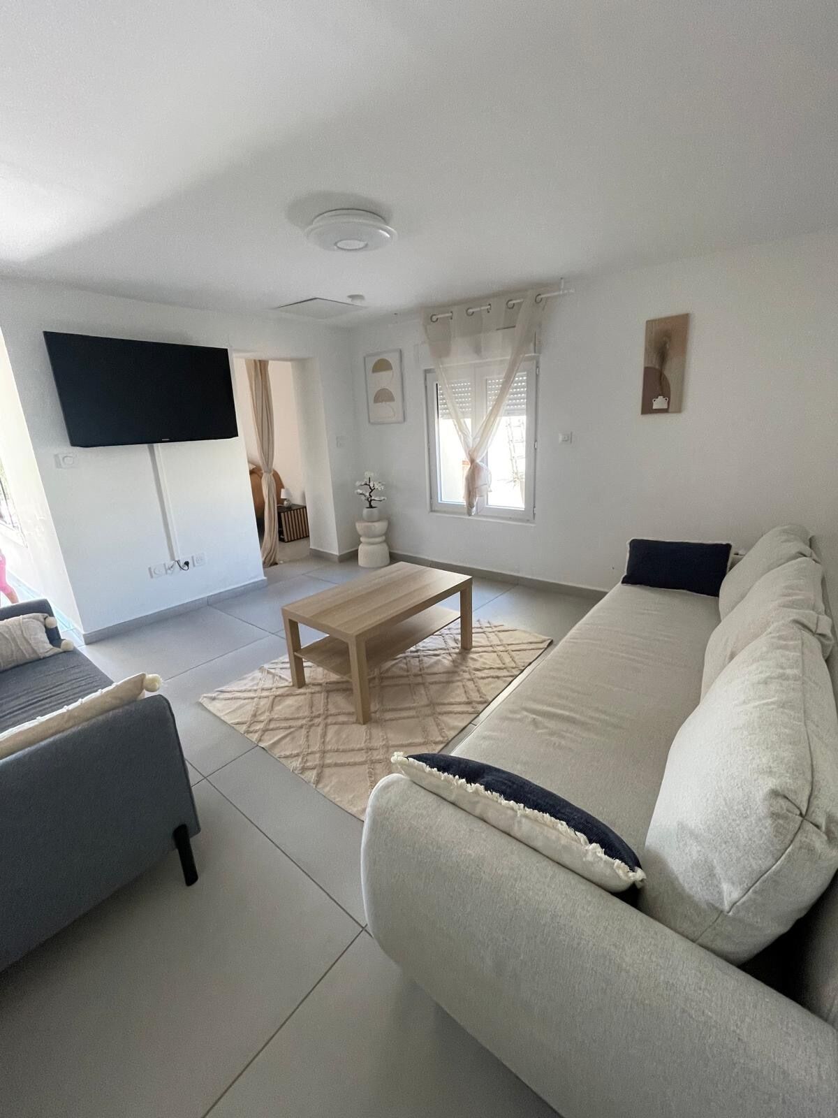 Una sala de estar con un sofá, una mesa de centro, un televisor montado en la pared y una ventana con cortinas.