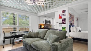 A modern living room with a grey sofa, a dining area with a table and chairs, a kitchen with a microwave, and a bedroom with a bed. - Music Themed BNB | Disco Lights | Fire Pit Lounge (Austin)