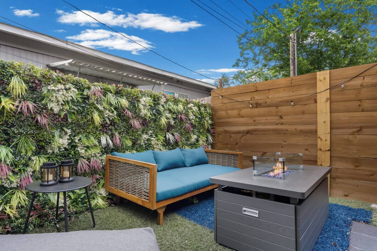 A patio area with a blue sofa, a small table with two lanterns, and a decorative wall with various plants.