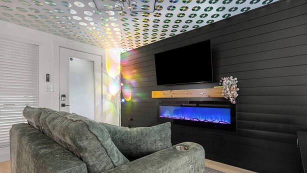 A modern living room with a grey sofa, a flat-screen TV mounted on a black wall, and a decorative fish tank. - Music Themed BNB | Disco Lights | Fire Pit Lounge (Austin)