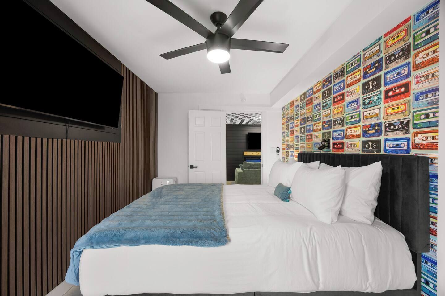Une chambre avec un lit, un ventilateur de plafond, un mur recouvert de cassettes audio colorées et un téléviseur fixé au mur.