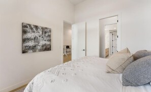 Een slaapkamer met een bed, kussens, een kunstwerk aan de muur en een deur naar een andere kamer.