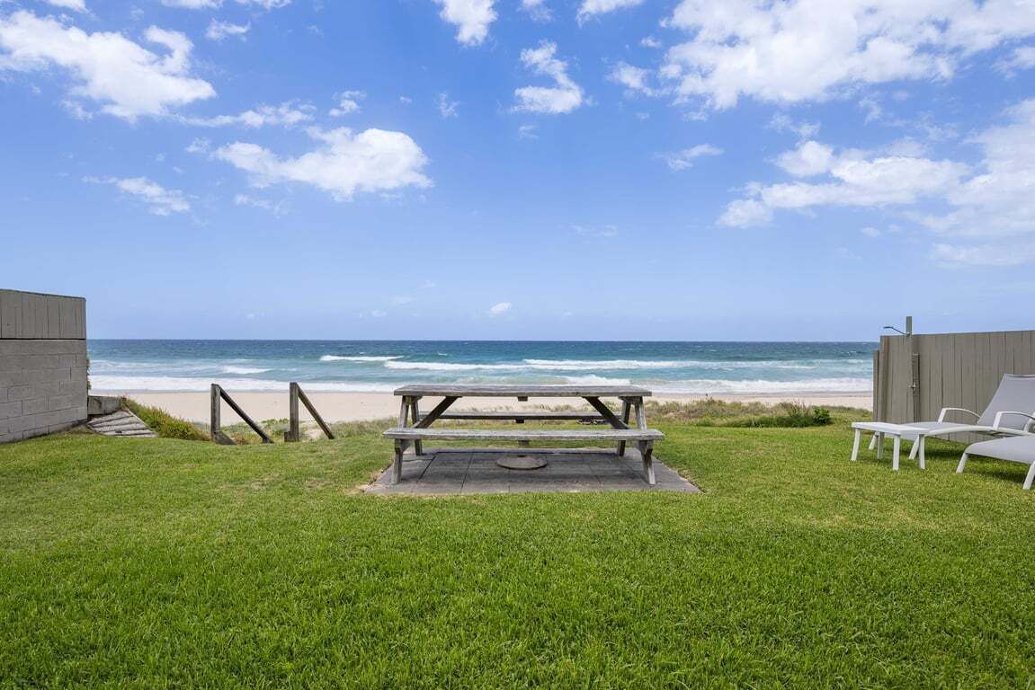 Ein grünes Areal im Freien mit einem Picknicktisch und weißen Lounge-Stühlen, mit Blick auf einen Strand mit Wellen und einem klaren Himmel.