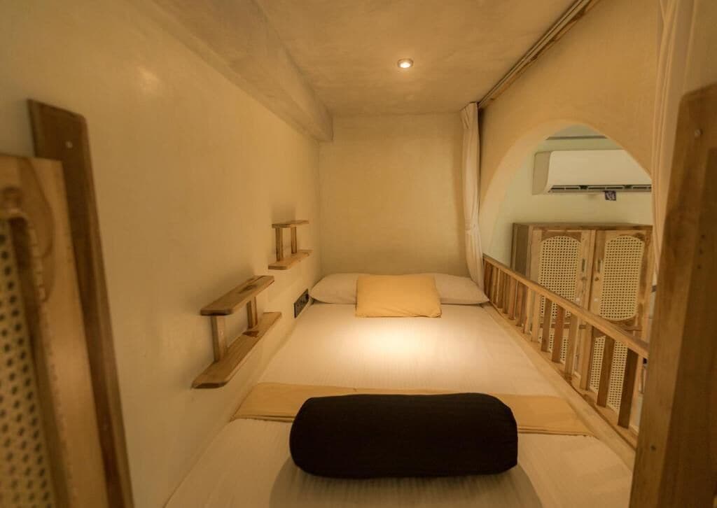Quarto de hotel pequeno e estreito, com uma cama, um pequeno banco de madeira, uma prateleira e uma janela com moldura de madeira.