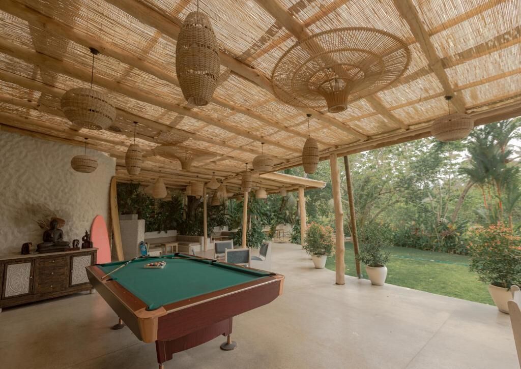 Un área de piscina interior con mesa de billar verde, lámparas de techo de mimbre y vista al jardín circundante.