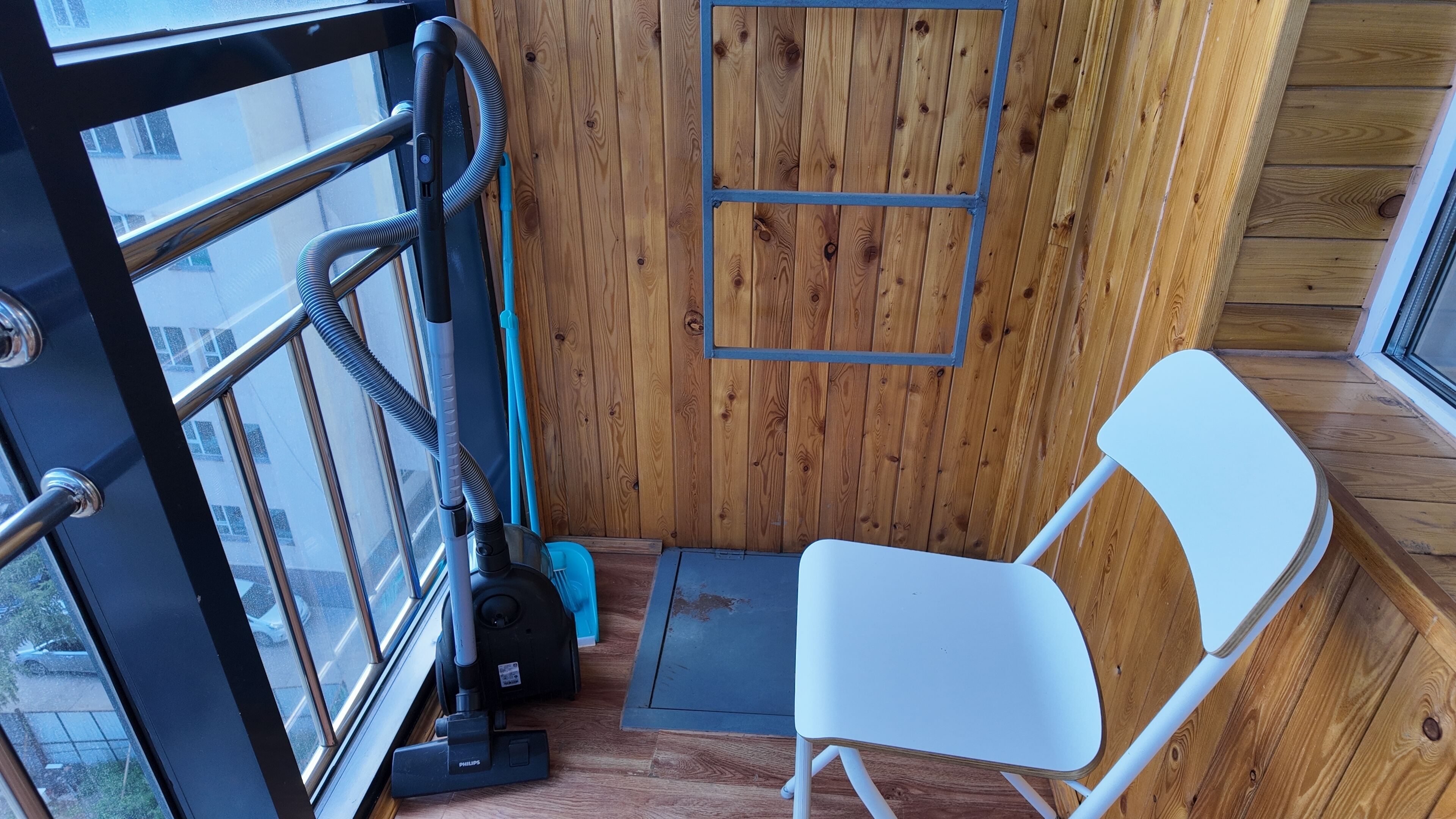 Een kleine kamer met een houten wand, een raam, een witte stoel en een stofzuiger.
