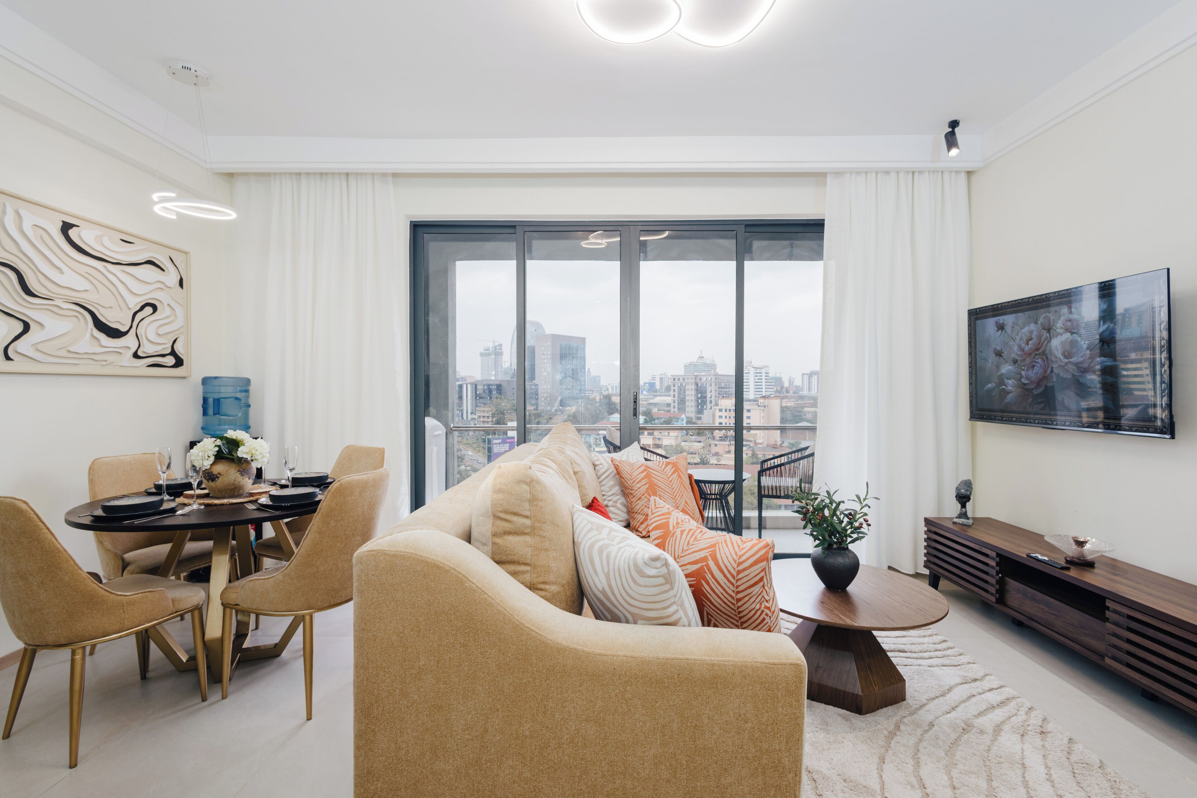 Un salon moderne avec vue sur la ville, un coin repas avec une table dressée pour deux personnes et un grand téléviseur fixé au mur.