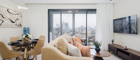 Un salon moderne avec vue sur la ville, un coin repas avec une table dressée pour deux personnes et un grand téléviseur fixé au mur.