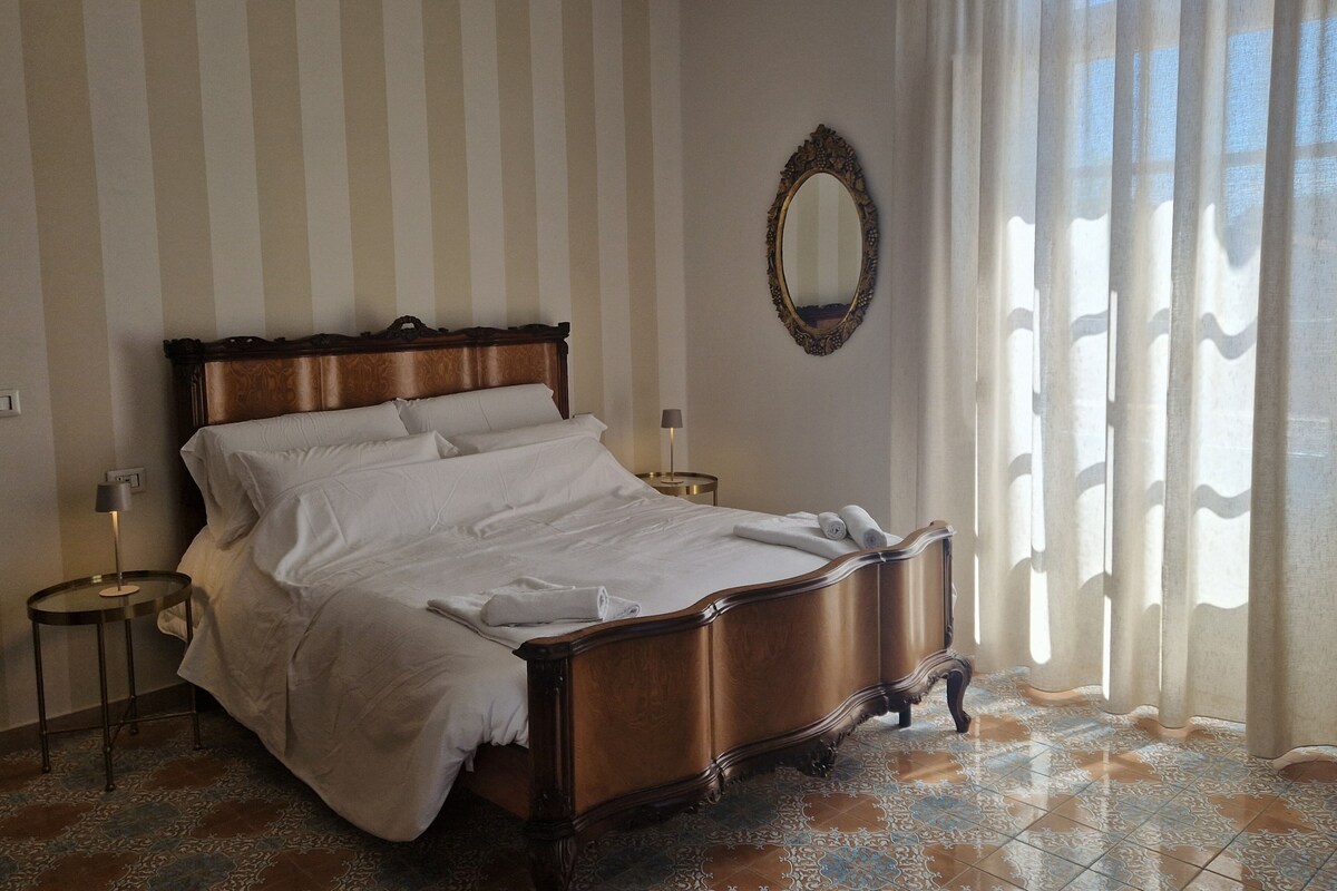 A hotel room with a large bed, a round mirror, and a view through sheer curtains.