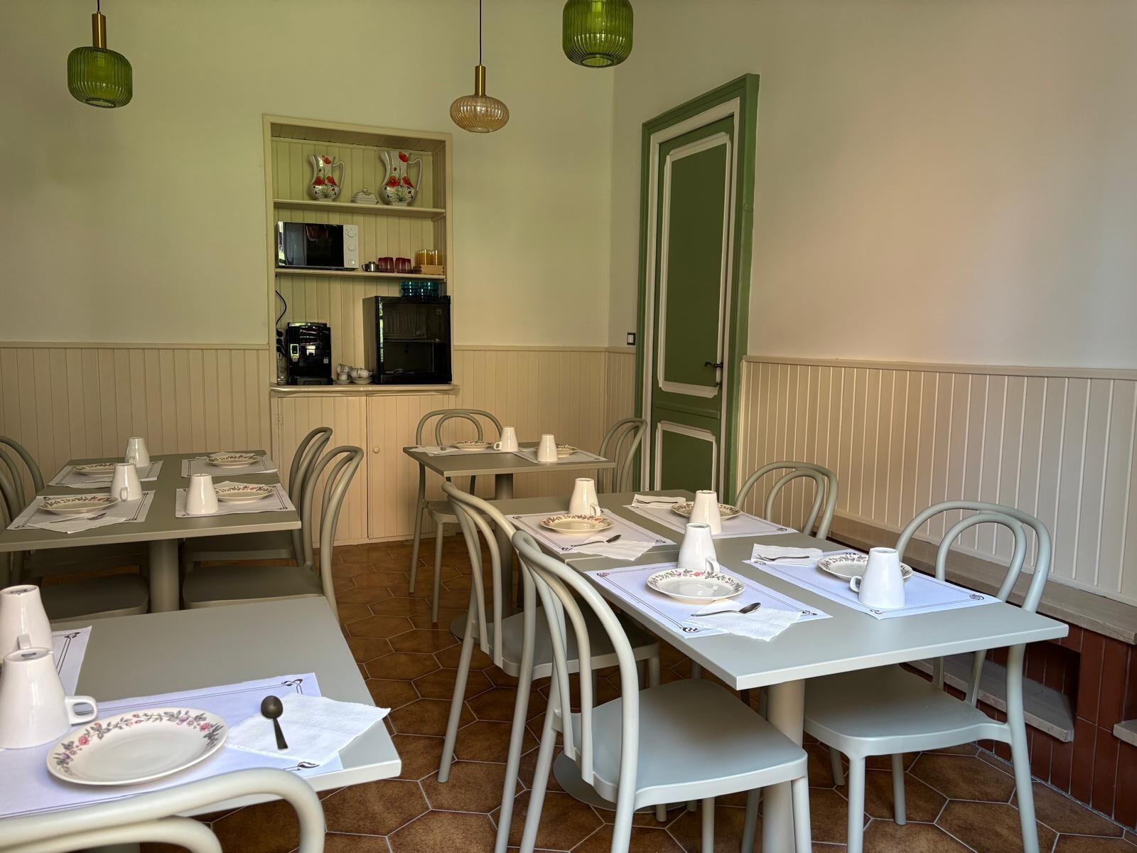 Comedor con mesas dispuestas para cuatro, sillas, zona de cocina con electrodomésticos y una puerta verde.
