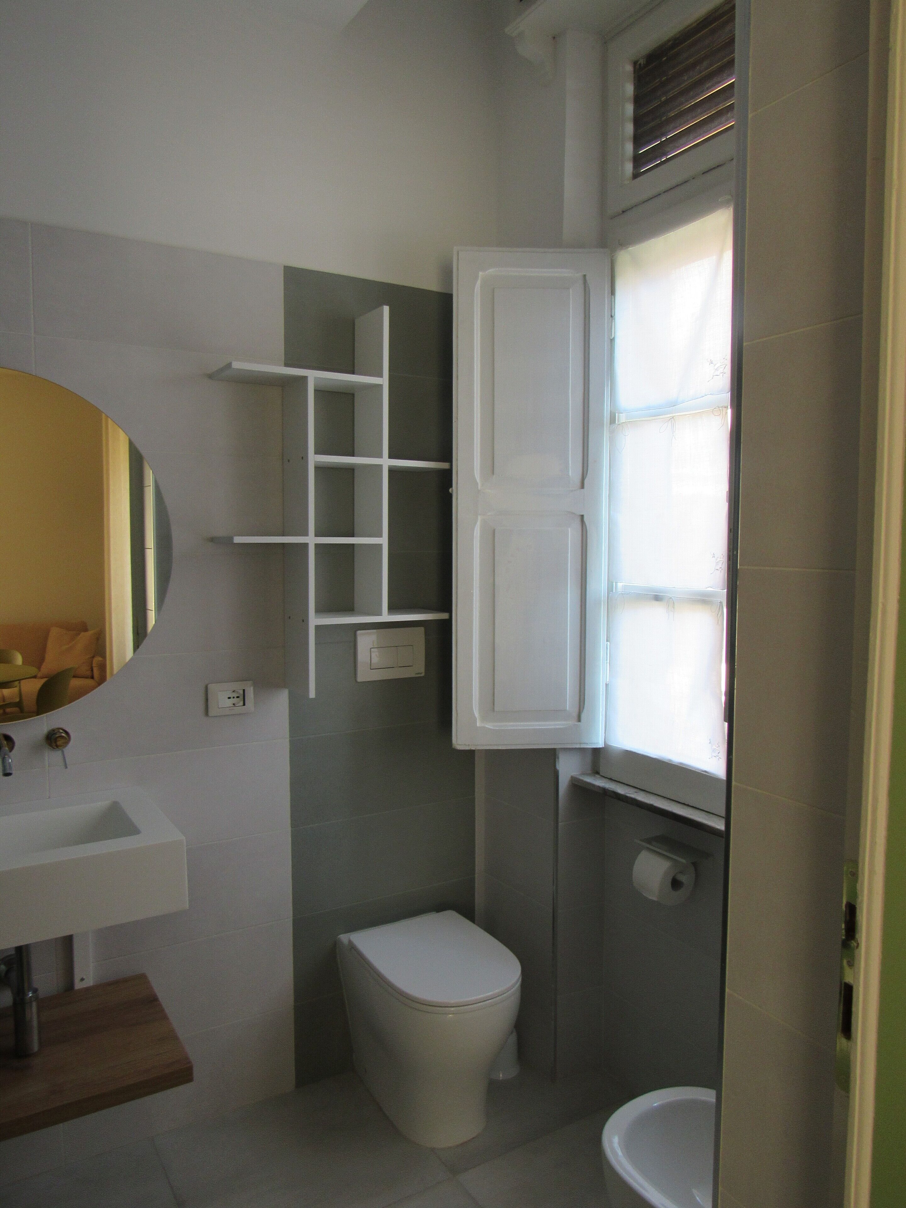 Ein Badezimmer mit Toilette, Waschbecken und Spiegel. Ein Fenster mit Jalousien und ein Ablagefach mit Pflegeprodukten.