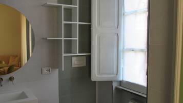 Ein Badezimmer mit Toilette, Waschbecken und Spiegel. Ein Fenster mit Jalousien und ein Ablagefach mit Pflegeprodukten.