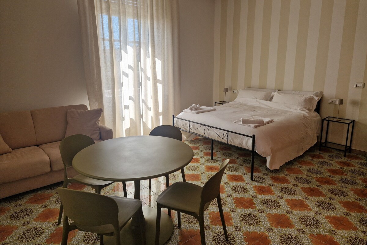 A hotel room with a bed, a sofa, a dining table with chairs, and a patterned floor.