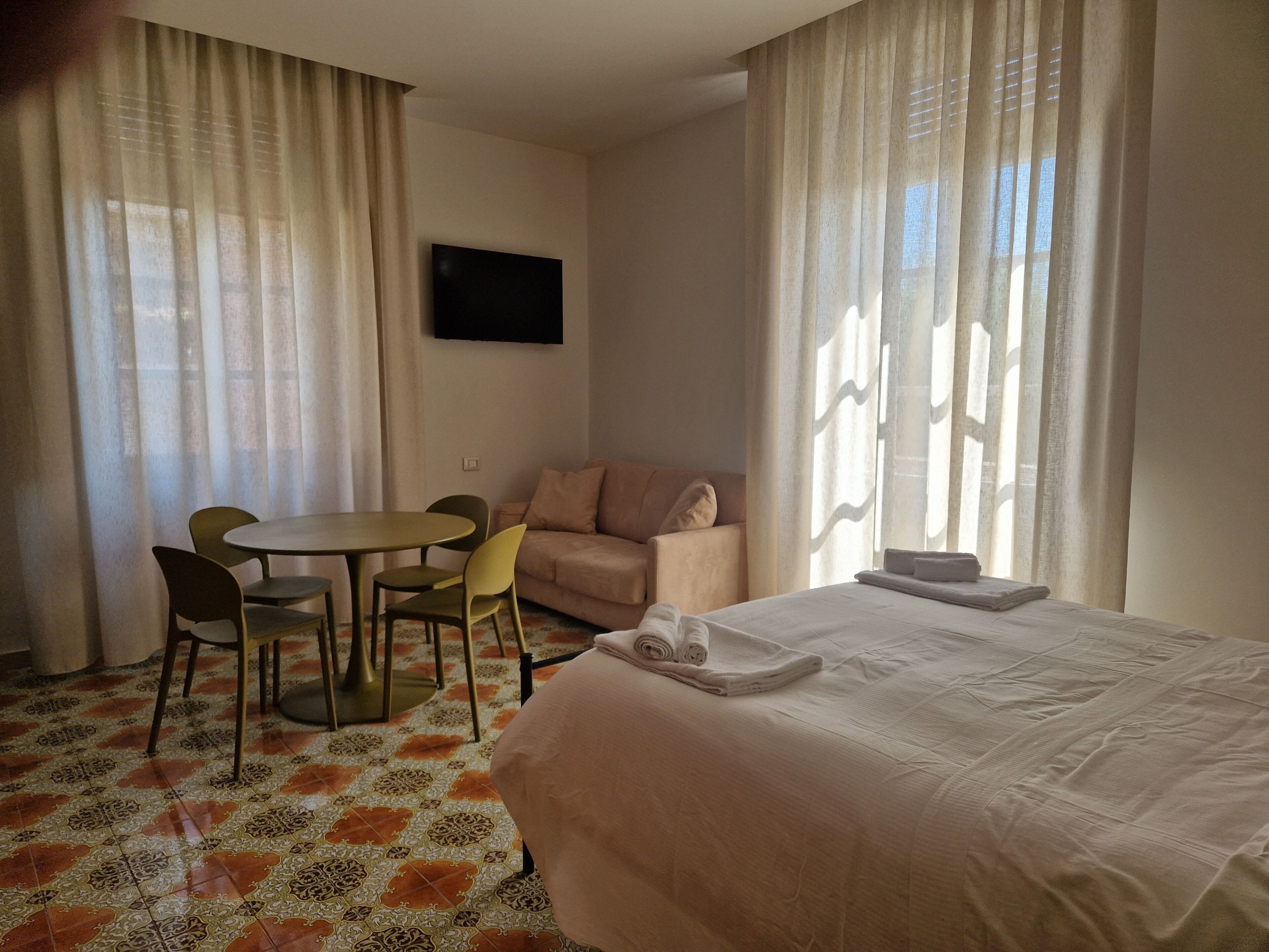 A hotel room with a bed, a sofa, a dining table with chairs, and a TV on the wall.