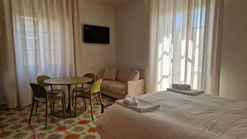 A hotel room with a bed, a sofa, a dining table with chairs, and a TV on the wall.