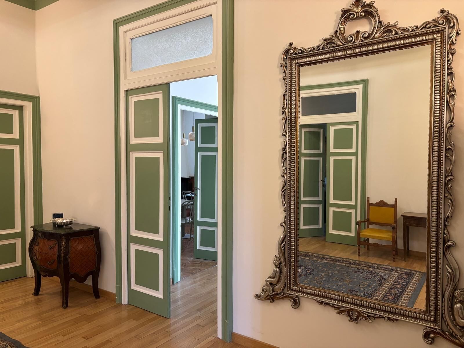 Una habitación con un espejo grande y decorativo, una mesita de madera y una puerta que da a otra estancia.