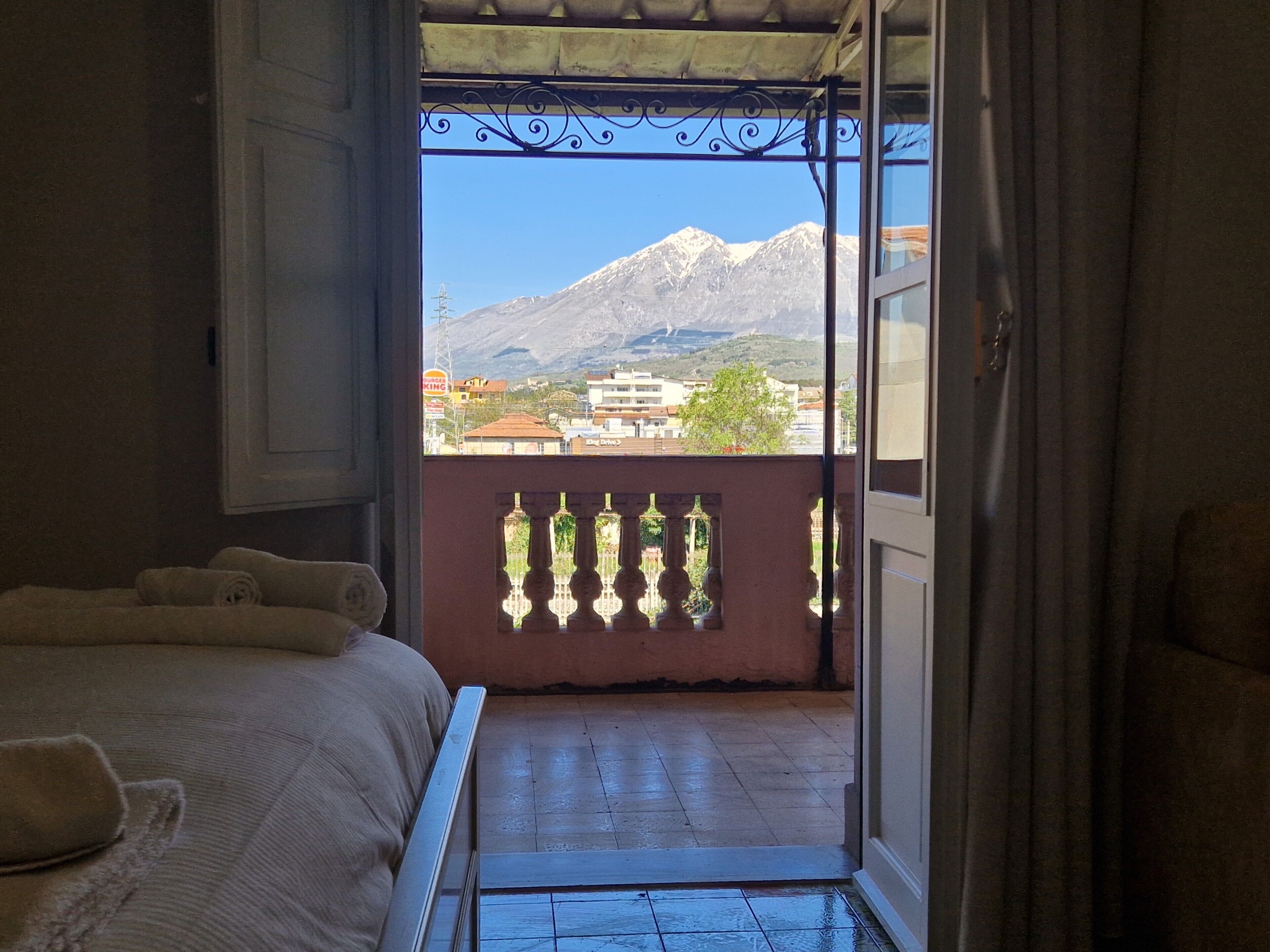 Ein Zimmer mit einem Bett, einem Balkon mit Blick auf Berge und Gebäude sowie einem Fenster mit einem dekorativen Eisengitter.
