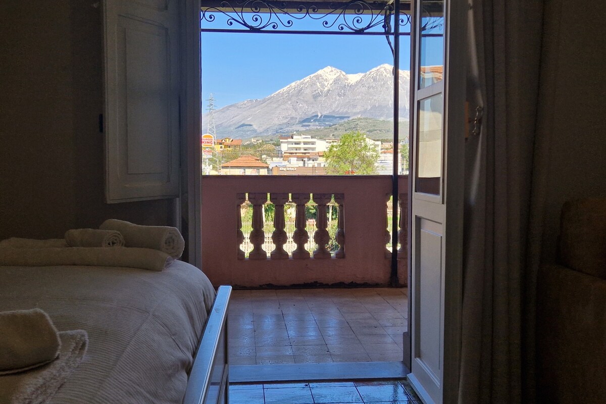 A room with a bed, a balcony with a view of mountains and buildings, and a window with a decorative iron grille.