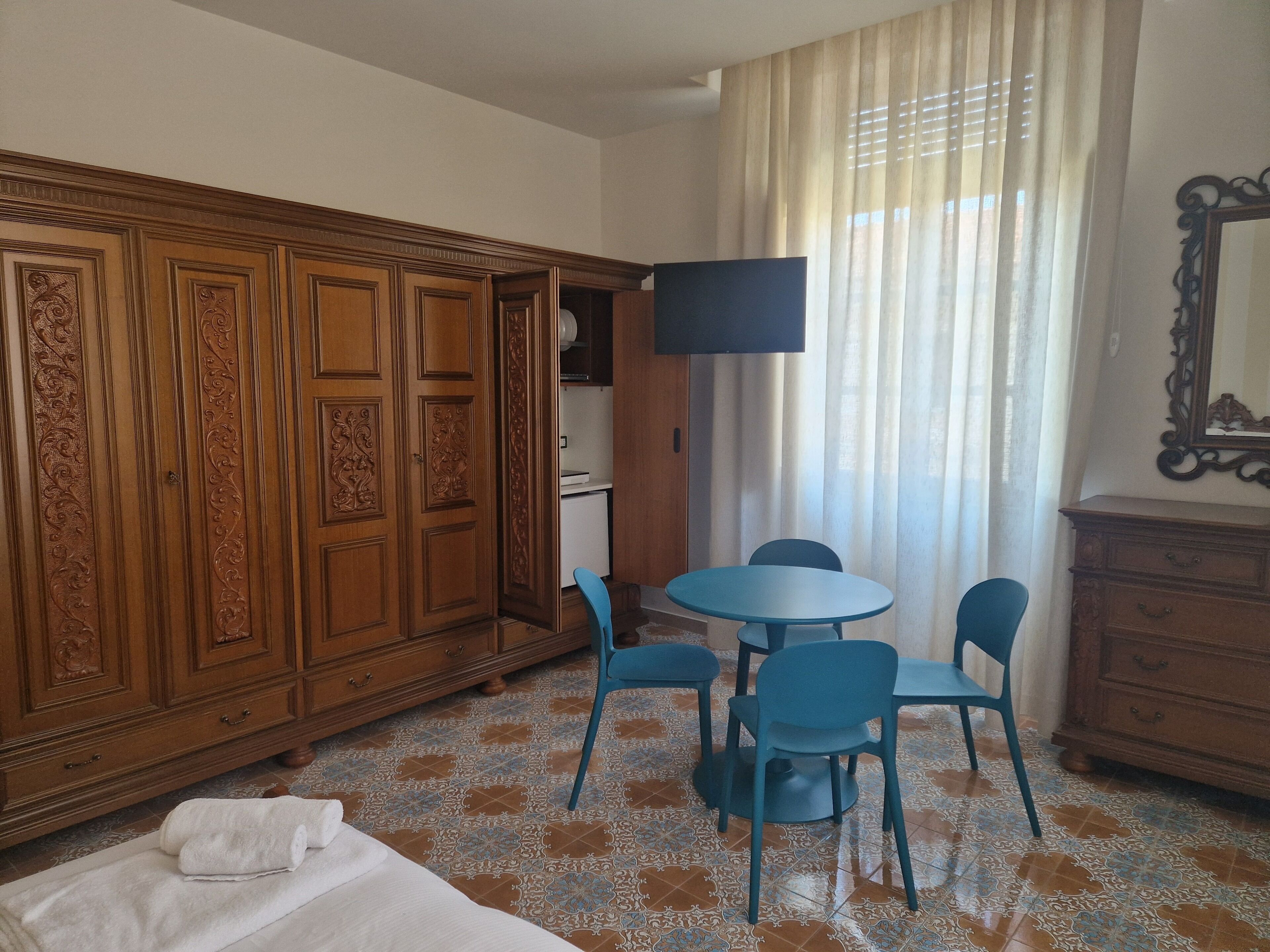 Una habitación de hotel con un armario de madera, una mesa redonda con sillas, un televisor montado en la pared y una cama con colcha blanca.