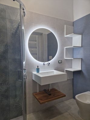 A modern bathroom with a glass shower enclosure, a white sink with a wooden shelf, and a round mirror with built-in lighting. - B&B il 900 (Avezzano)