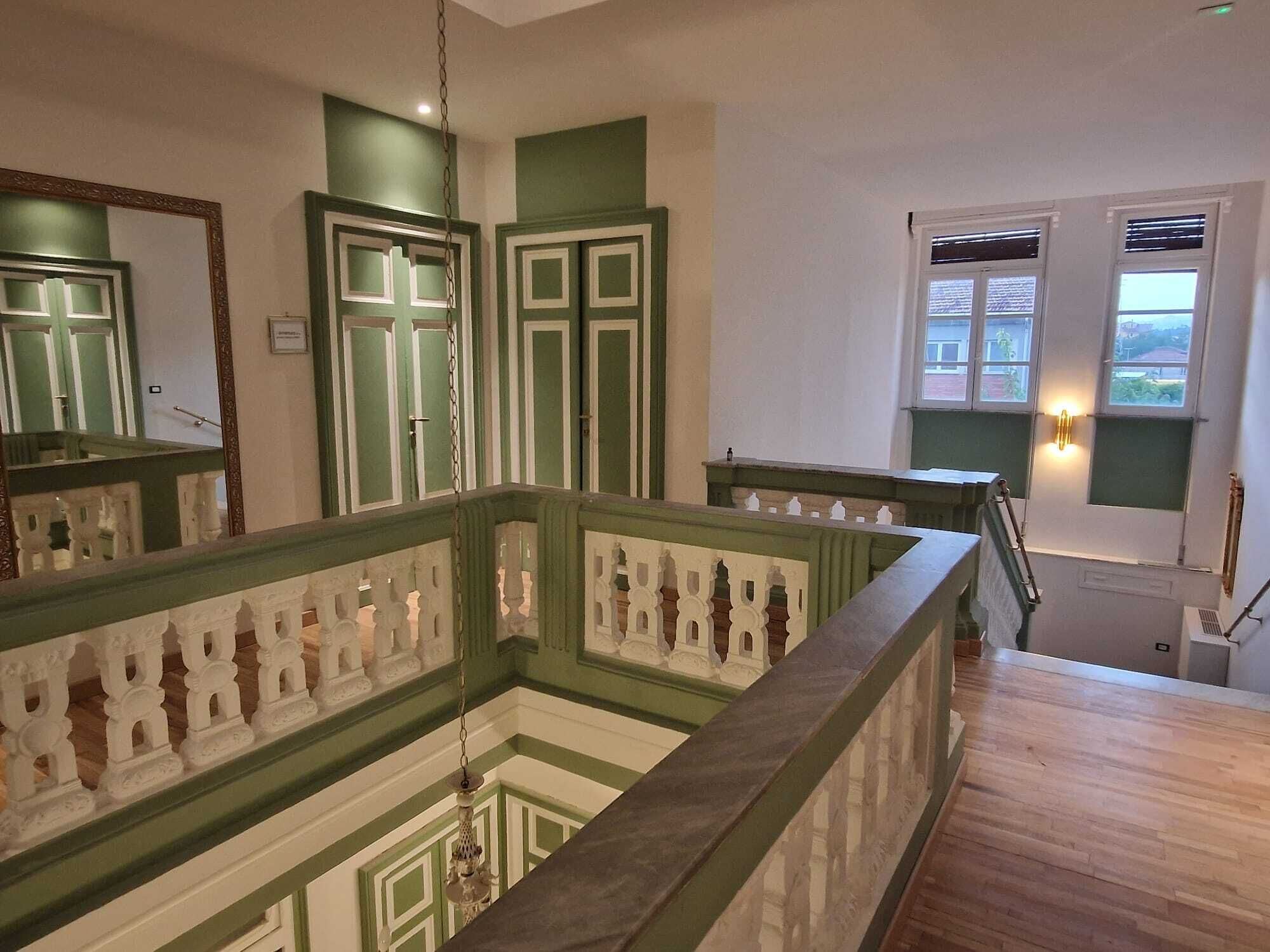 Una escalera con barandilla de madera y pasamanos verde, que conduce a un rellano con vistas al exterior.