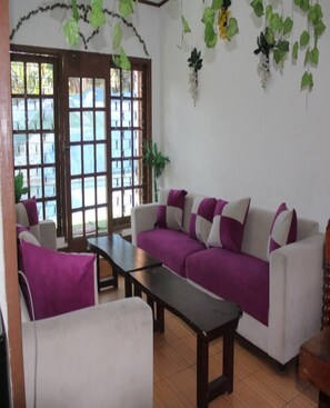 A living room with a sofa, a coffee table, and a chair. There are windows with a view of a blue sky and greenery. - Cozzy Harys Farm Pangalengan (Pangalengan)