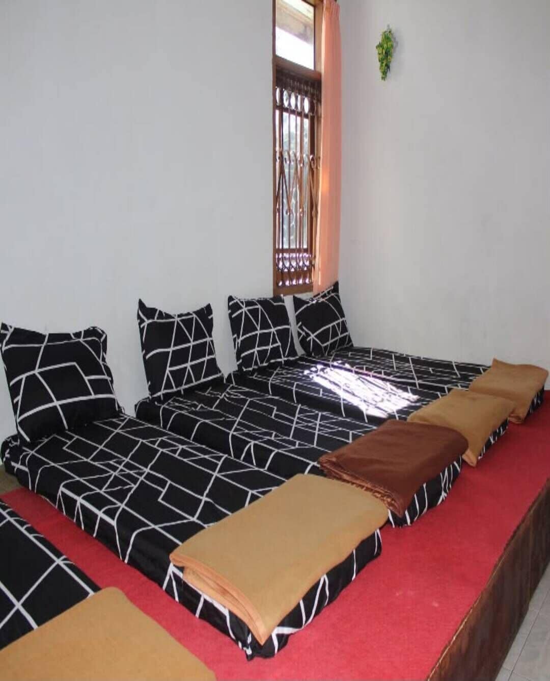 Una habitación con cama, almohadas, alfombra roja y ventana con cortina.