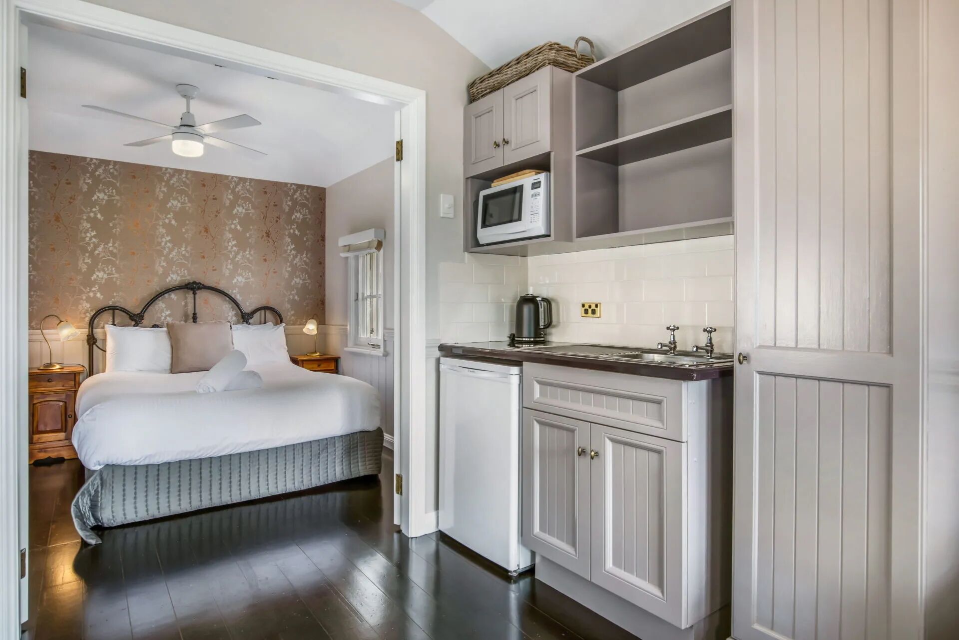 A bedroom with a bed, nightstand, lamp, and a ceiling fan. Adjacent is a kitchenette with a microwave, kettle, and cabinets.