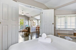 A hotel room with a bed, a desk, a chair, a television, and a window with blinds. - Violet by Hyams Beach Seaside Cottages | Romantic, iconic heritage cottage (Hyams Beach)