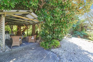 A covered outdoor seating area with wicker furniture, surrounded by lush greenery and a gravel path. - Violet by Hyams Beach Seaside Cottages | Romantic, iconic heritage cottage (Hyams Beach)