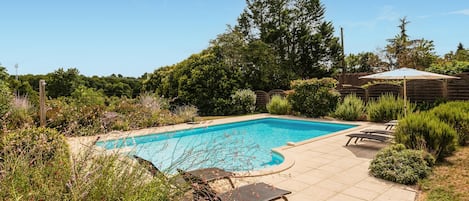 A rectangular swimming pool with a tiled deck, surrounded by lush greenery and a wooden fence.